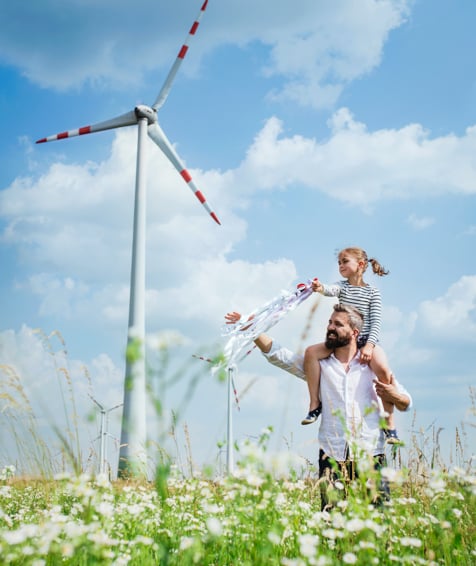 An individual and their child walking in a field next to a wind turbine.